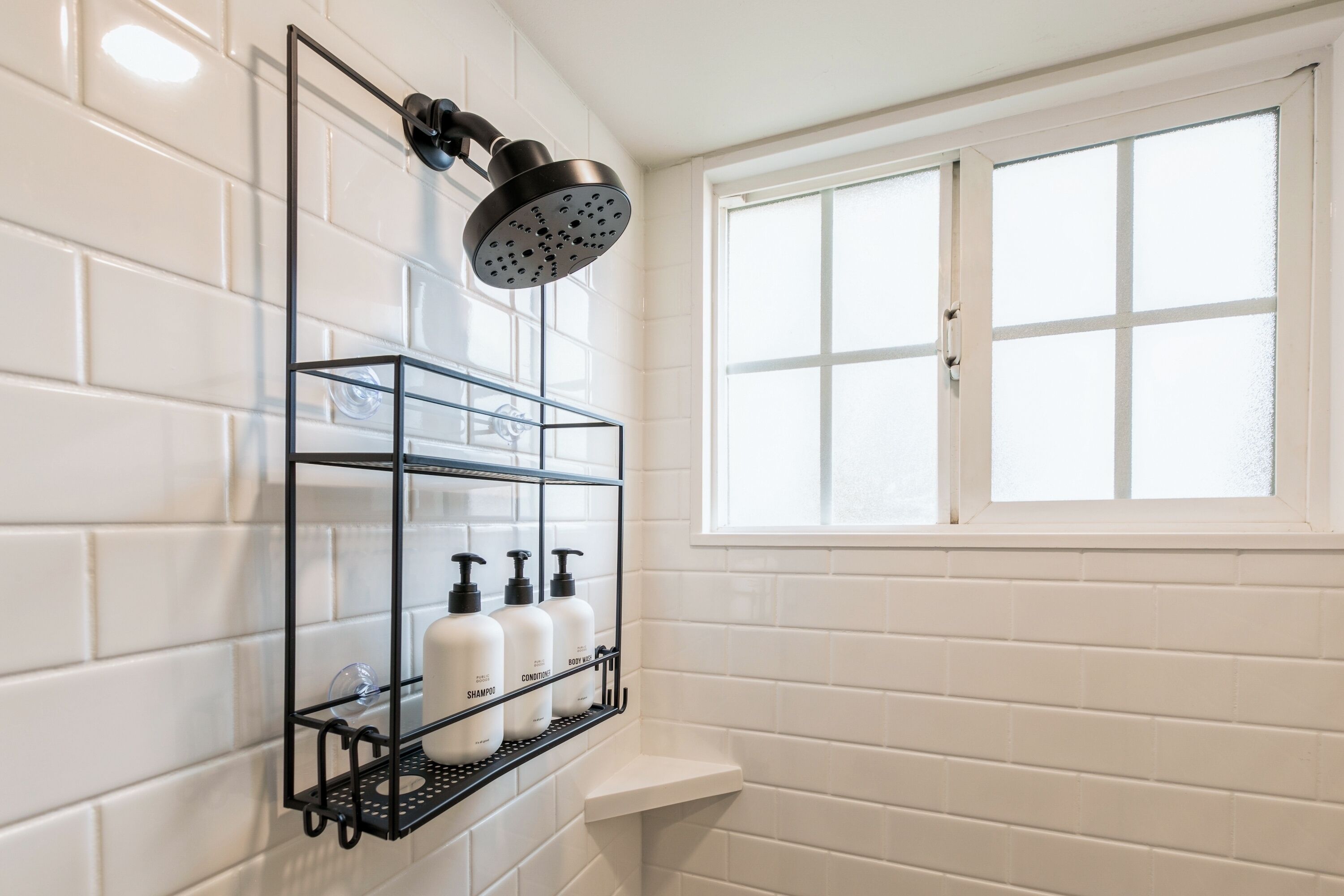 A bathroom with white tiled walls, a window, a shower with a black head, and a shelf with toiletries.