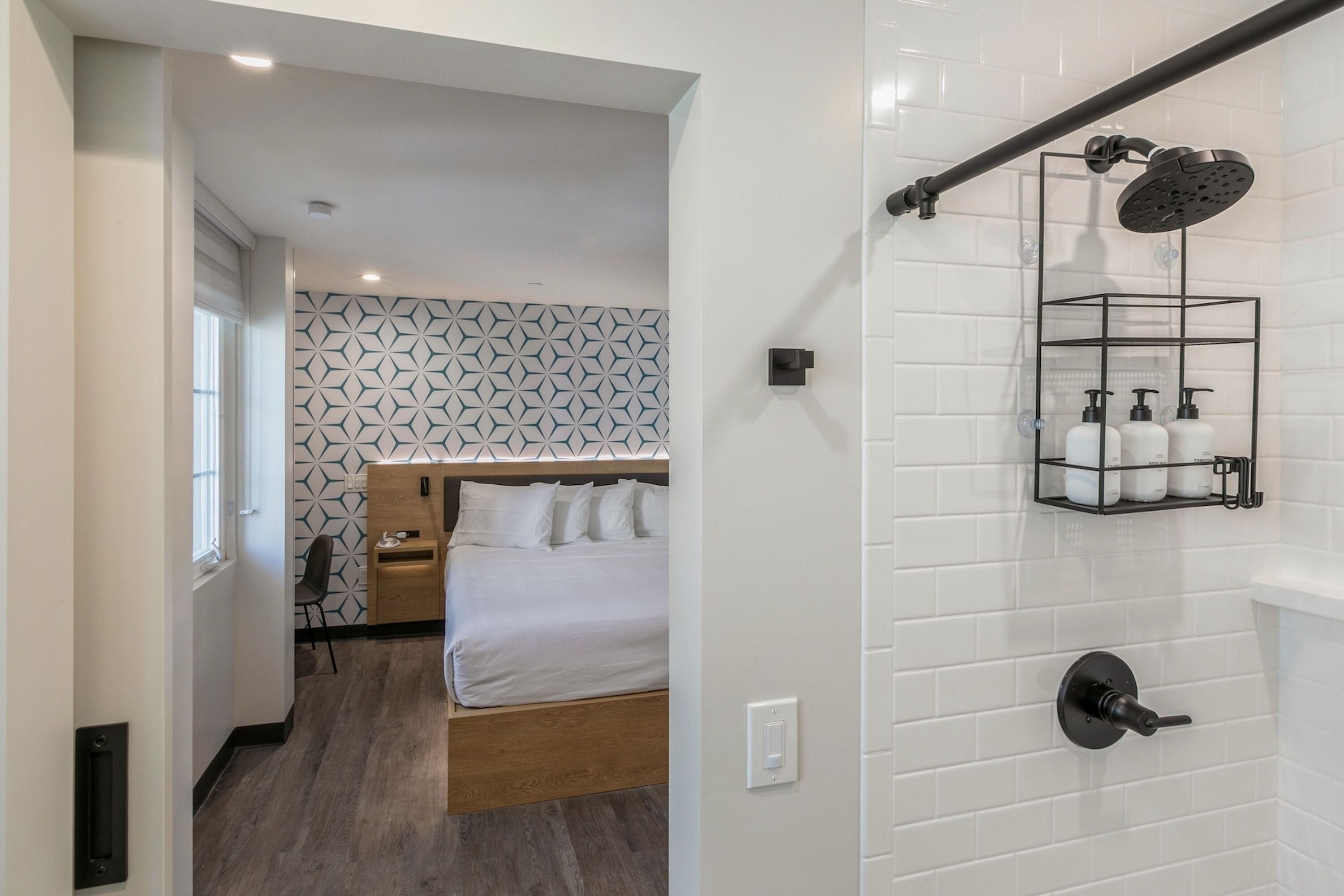 Habitación de hotel moderna con cama de madera, ducha con manguera negra y pared de azulejos blancos con un diseño geométrico.