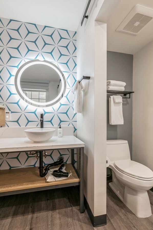 Un baño moderno con un muro de diseño geométrico, un espejo redondo, un lavabo blanco y un inodoro.