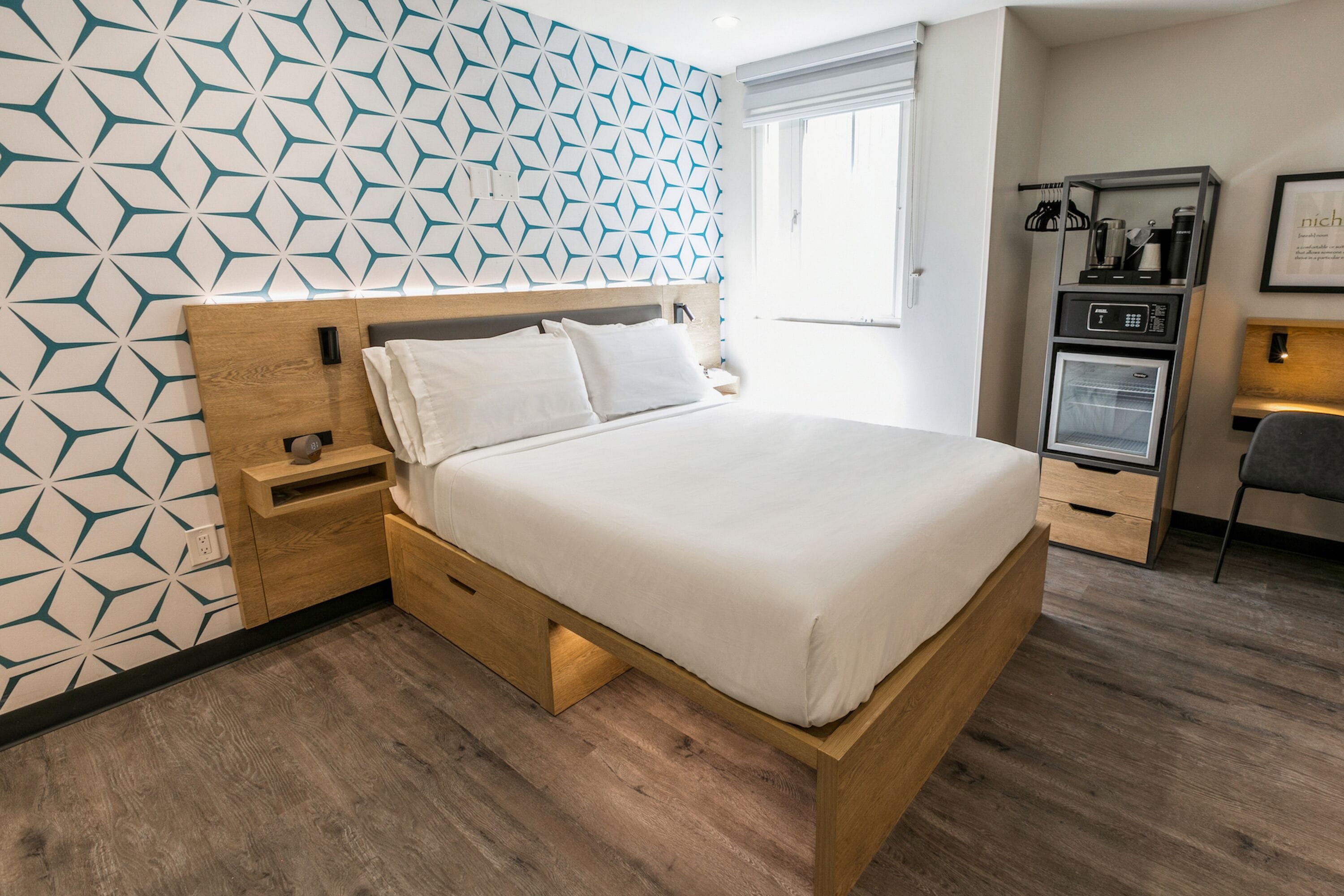 A modern hotel room with a geometric patterned wall, a wooden bed frame, a bedside table, and a built-in mini-fridge.