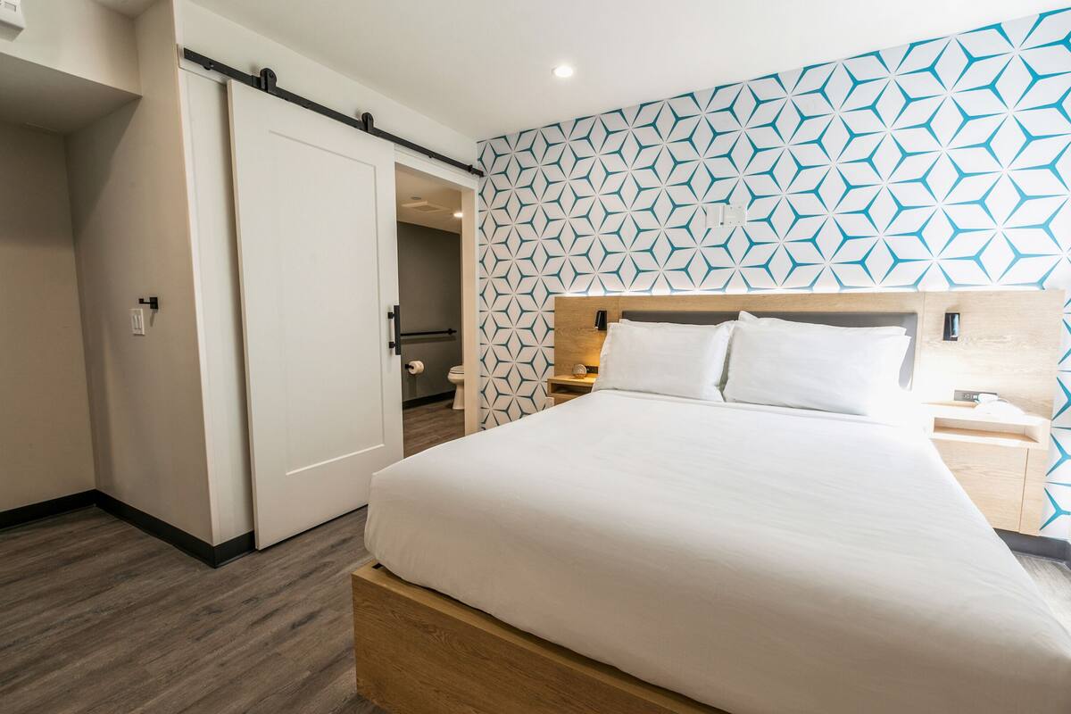 A hotel room with a bed, a nightstand, a wooden headboard, a geometric patterned wall, and a door leading to a bathroom.