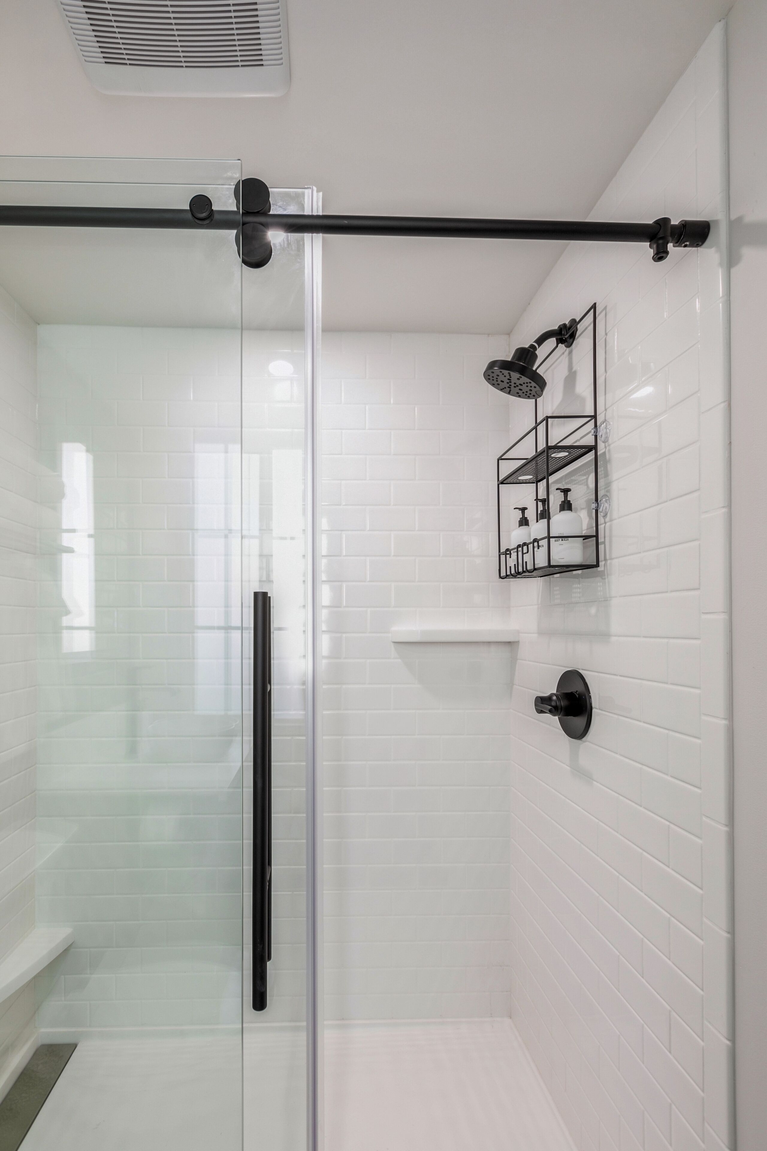 A modern bathroom with white tiled walls, a glass shower enclosure, a black towel rack, and a wall-mounted soap dispenser.