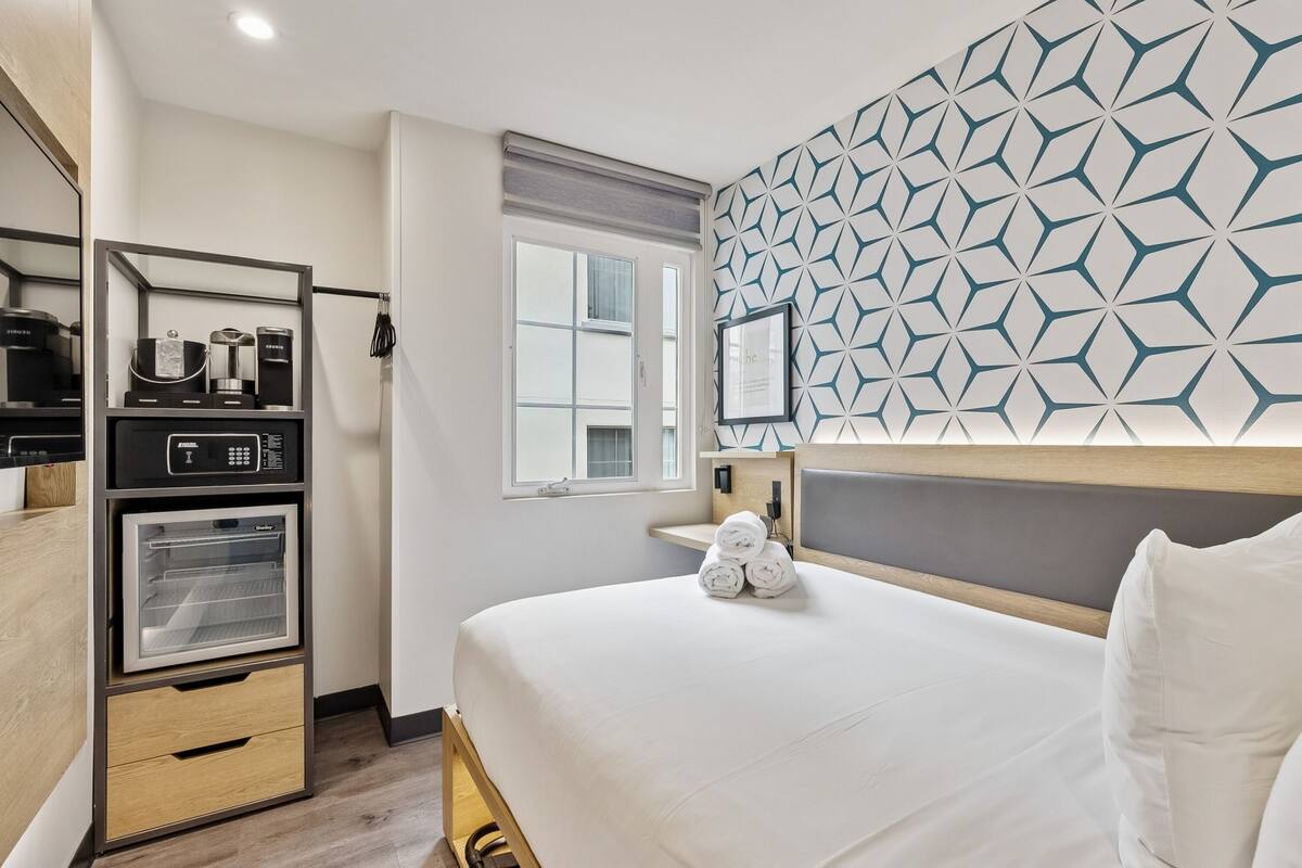 A hotel room with a bed, a built-in coffee machine, a mini-fridge, and a geometric-patterned wall.