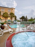 Piscina con sillas de descanso y jacuzzi, rodeada de palmeras y un edificio con balcones.