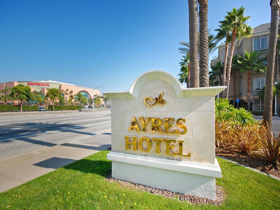 A hotel sign with "AYRES HOTEL" written in gold letters, surrounded by palm trees and a clear blue sky.