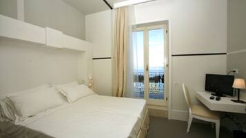 Quarto de hotel com uma cama, uma escrivaninha com computador e vista para o mar pela janela.