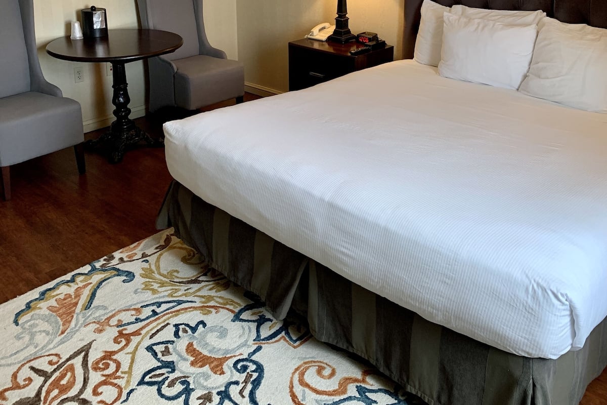 A hotel room with a large bed, two armchairs, a small table, a lamp, and a patterned rug.