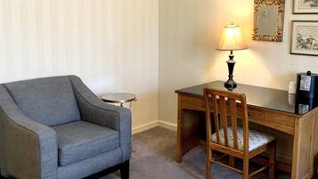 Una habitación de hotel con un sillón gris, un escritorio de madera con una lámpara y una mesa redonda pequeña.