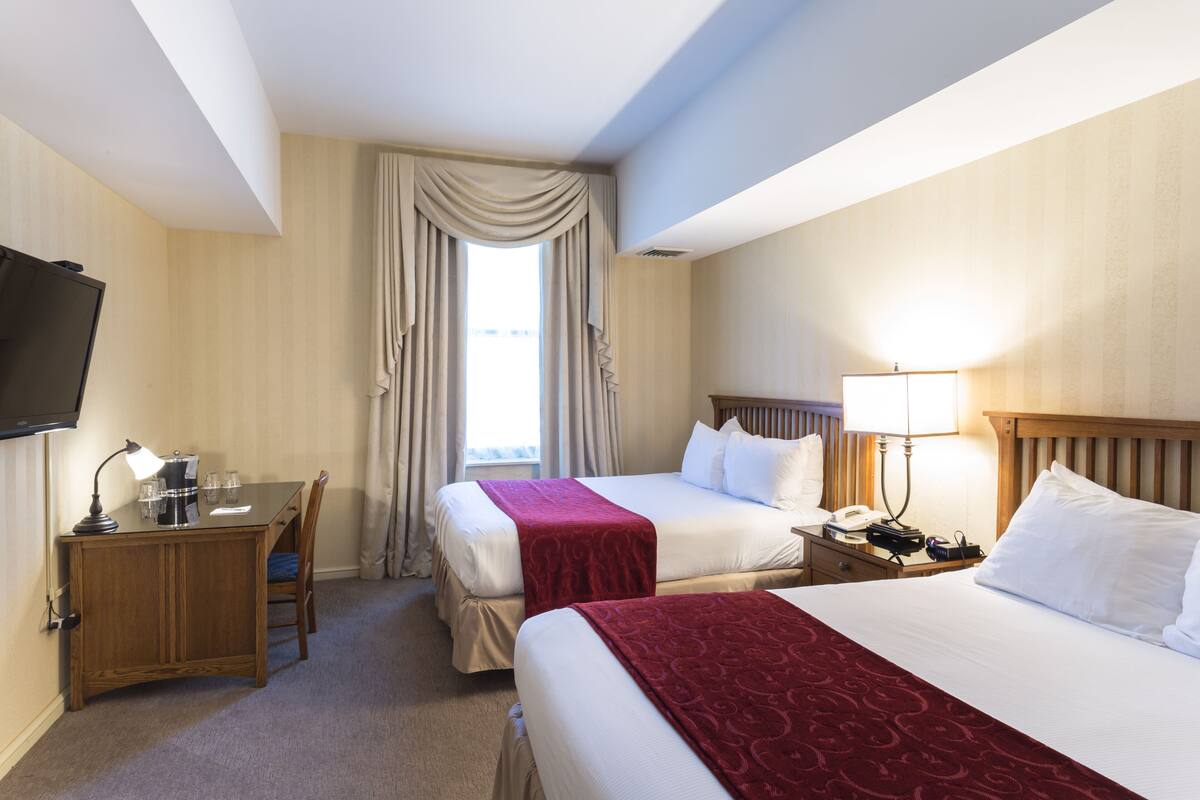 A hotel room with two beds, a desk with a lamp, a television, and a window with curtains.