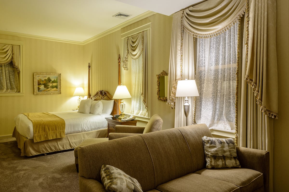 A hotel room with a bed, two bedside tables with lamps, a sofa, and a large window with curtains.