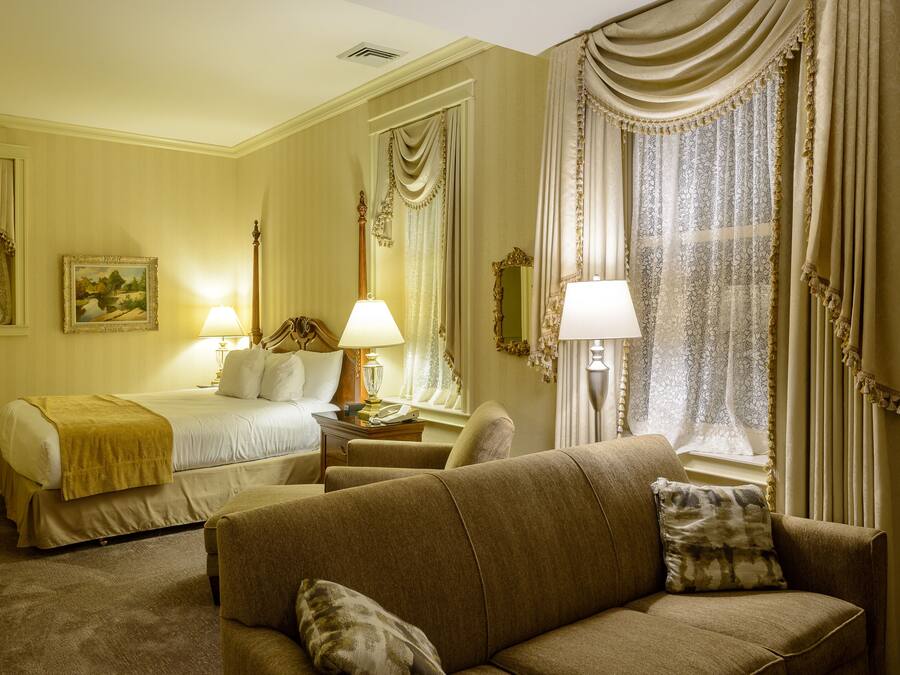 Une chambre d’hôtel avec un lit, deux tables de chevet avec des lampes, un canapé et une grande fenêtre avec des rideaux.