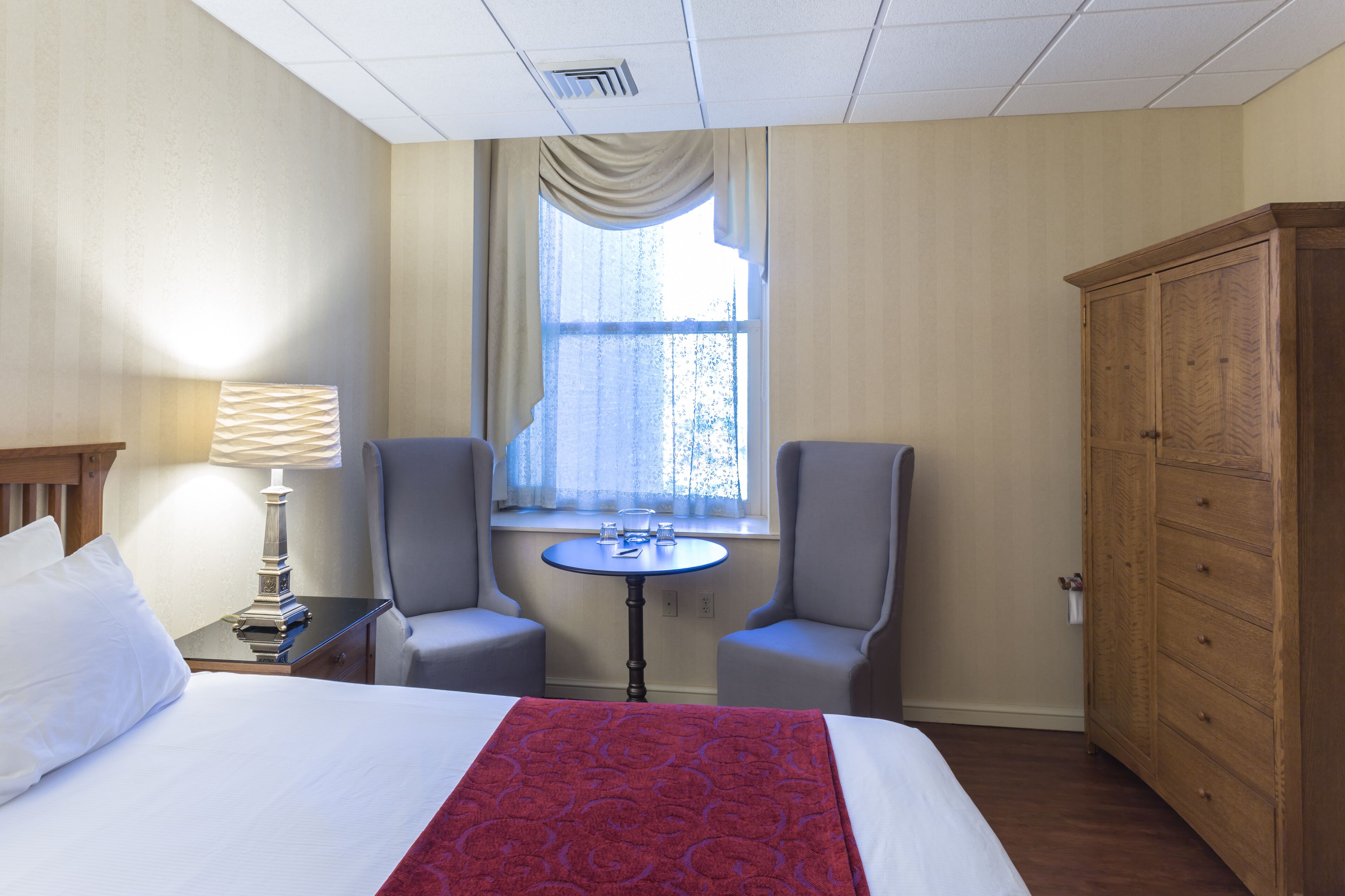 A hotel room with a bed, two armchairs, a small round table, a lamp, a wardrobe, and a window with curtains.