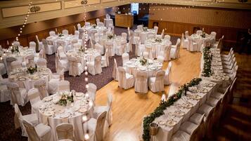 A banquet hall with round tables arranged in a semi-circle, each set with white tablecloths and chairs, and a long table with a floral arrangement down the center.