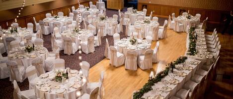 Un salón de banquetes con mesas redondas dispuestas en forma de semicírculo, cada una con mantel blanco y sillas, y una mesa larga con un arreglo floral en el centro.