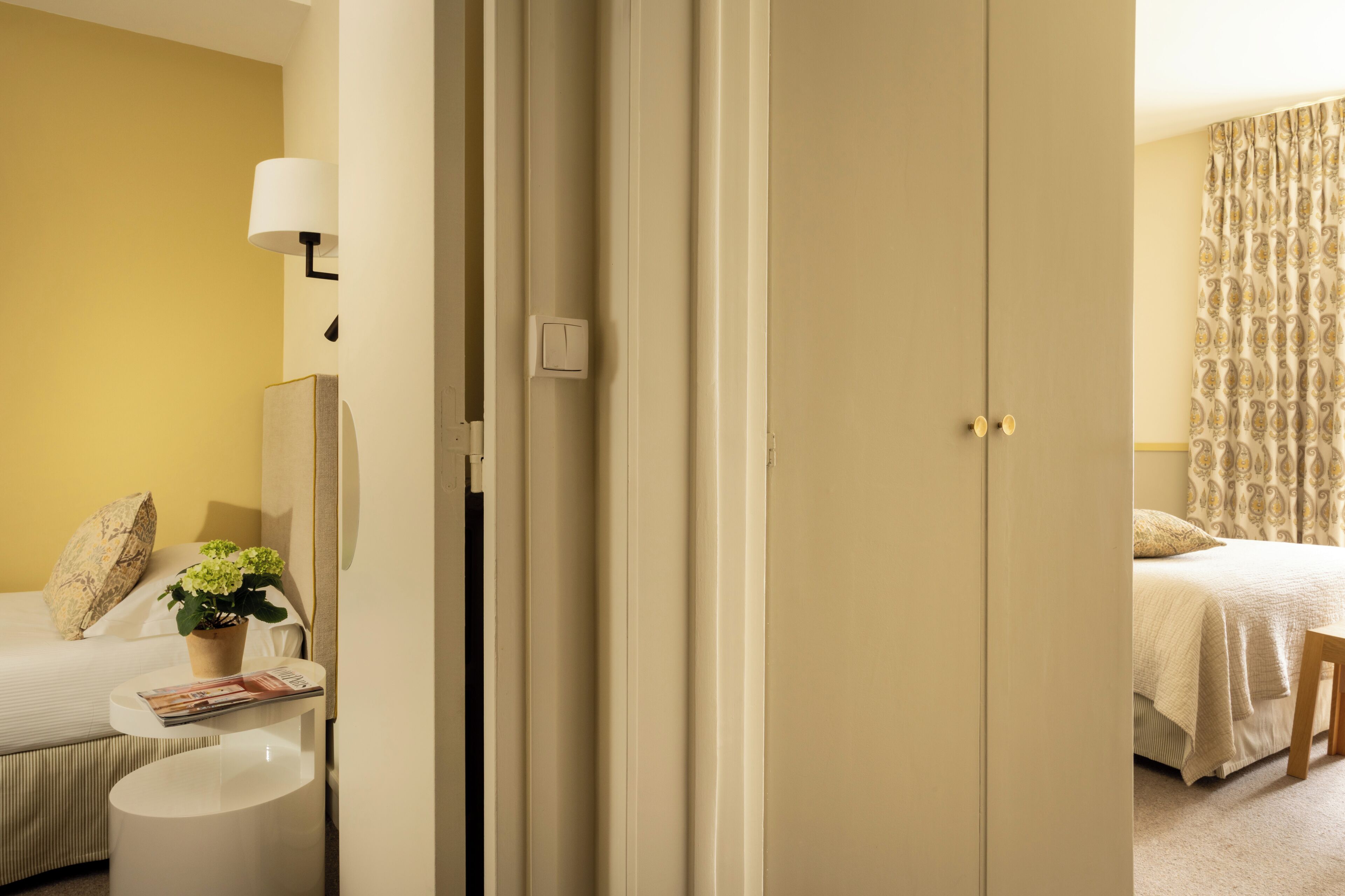 Une chambre d’hôtel avec un lit, une table de chevet avec une lampe, une plante en pot et une porte donnant sur une autre pièce.