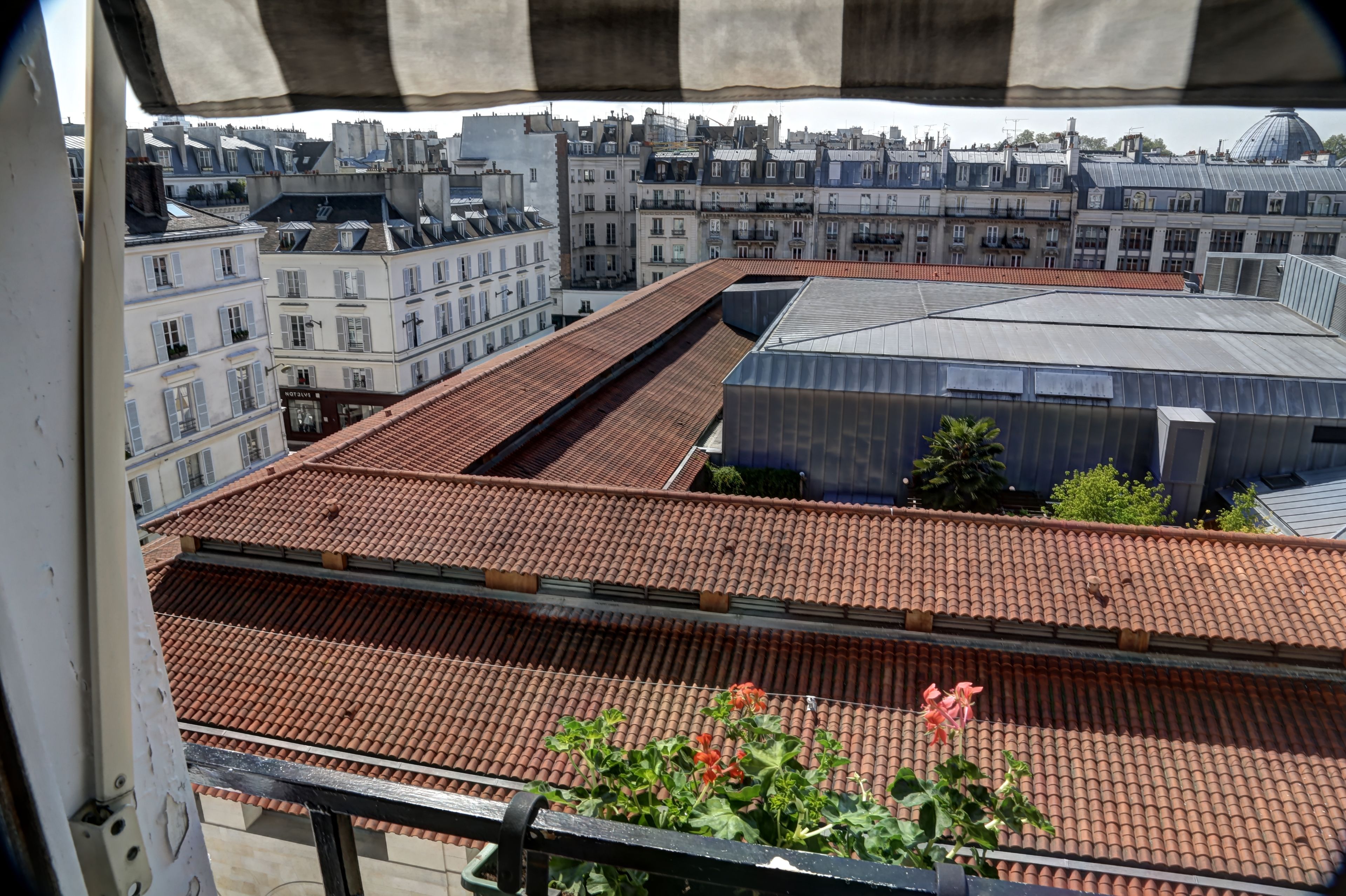 A view from a balcony with a tiled roof, overlooking a cityscape with various buildings, including a large industrial structure and residential buildings with multiple stories.