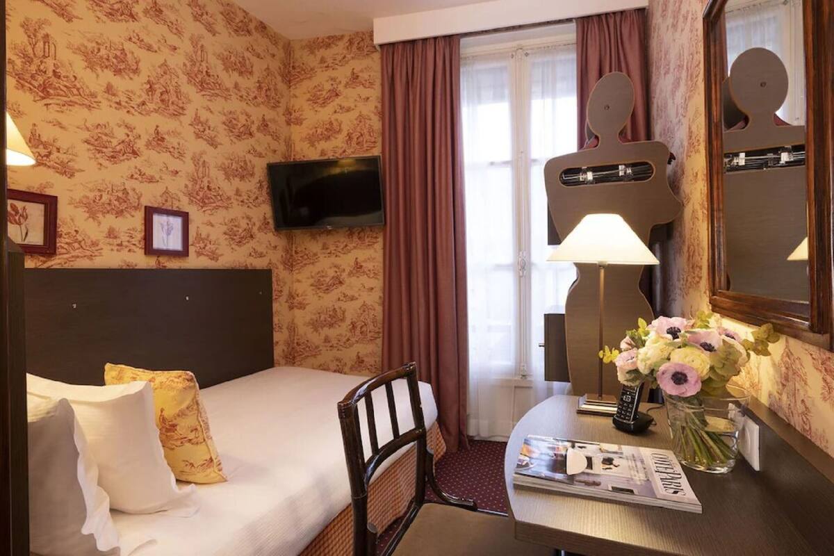 A hotel room with a bed, a desk with a lamp, a mirror, and a vase with flowers.