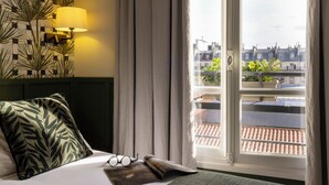 A hotel room with a bed, a lamp, a view of the city through a window, and a plant on the windowsill. - Hôtel Clément (Paris)