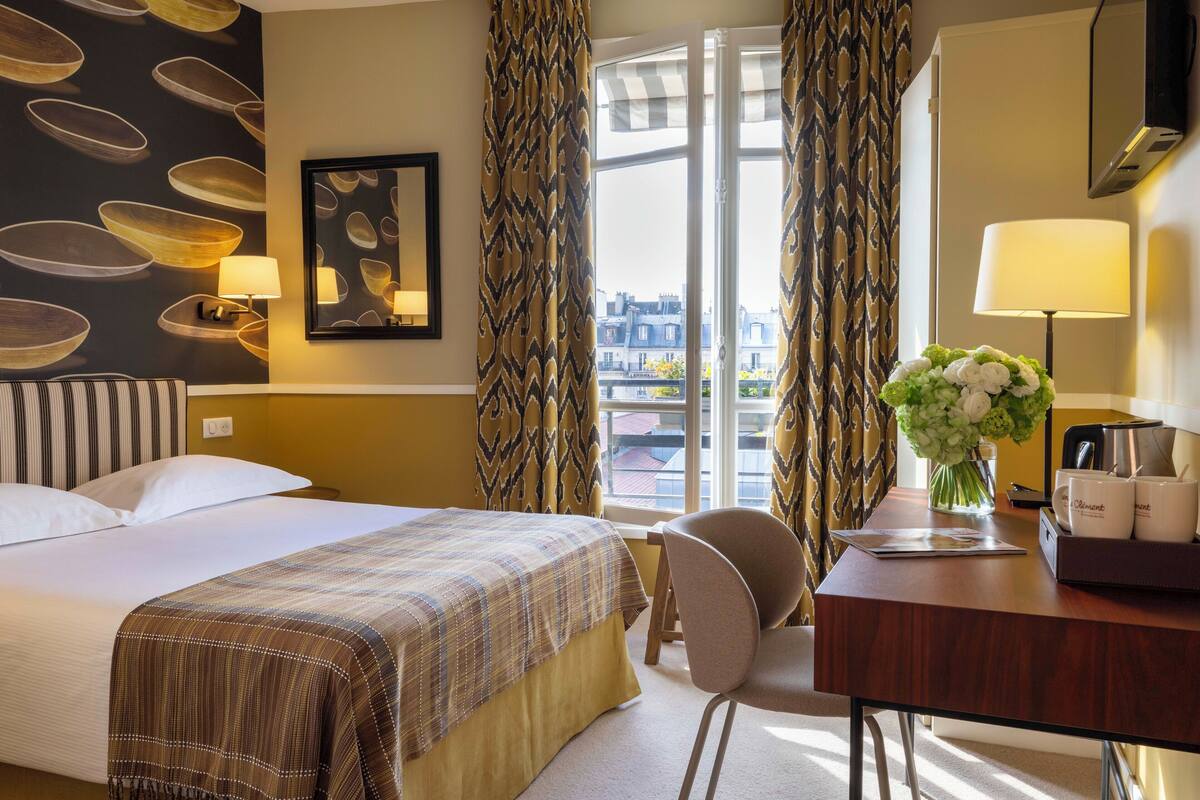 A hotel room with a bed, a desk with a chair, a coffee maker, and a vase with flowers.