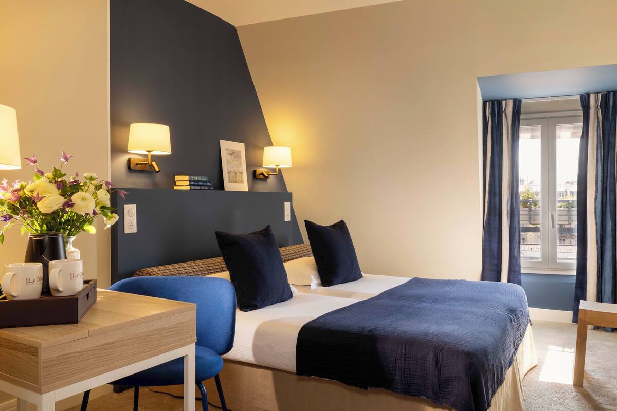 A hotel room with a bed, two bedside lamps, a desk with a vase of flowers, and a window with curtains.