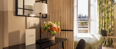 Habitación de hotel con cama, escritorio con lámpara, un jarrón de flores y vistas a edificios a través de la ventana, además de un letrero en la pared.