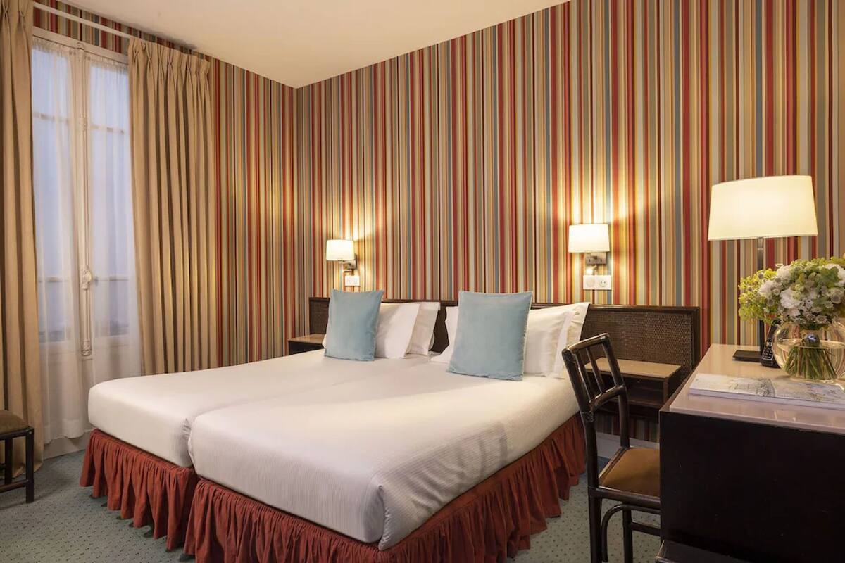 A hotel room with a large bed, two bedside tables with lamps, a desk with a chair, and a window with curtains.