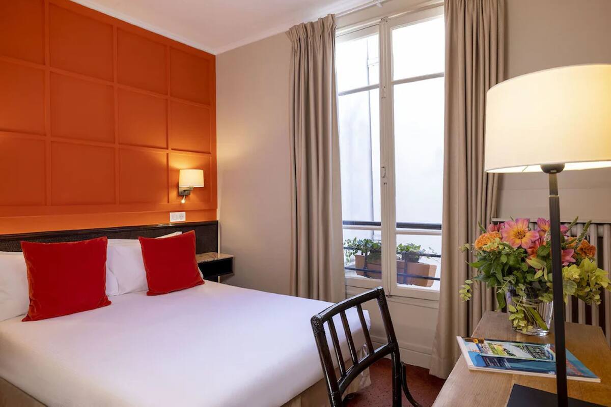 A hotel room with a bed, a chair, a table with a lamp, a window with curtains, and a vase with flowers.