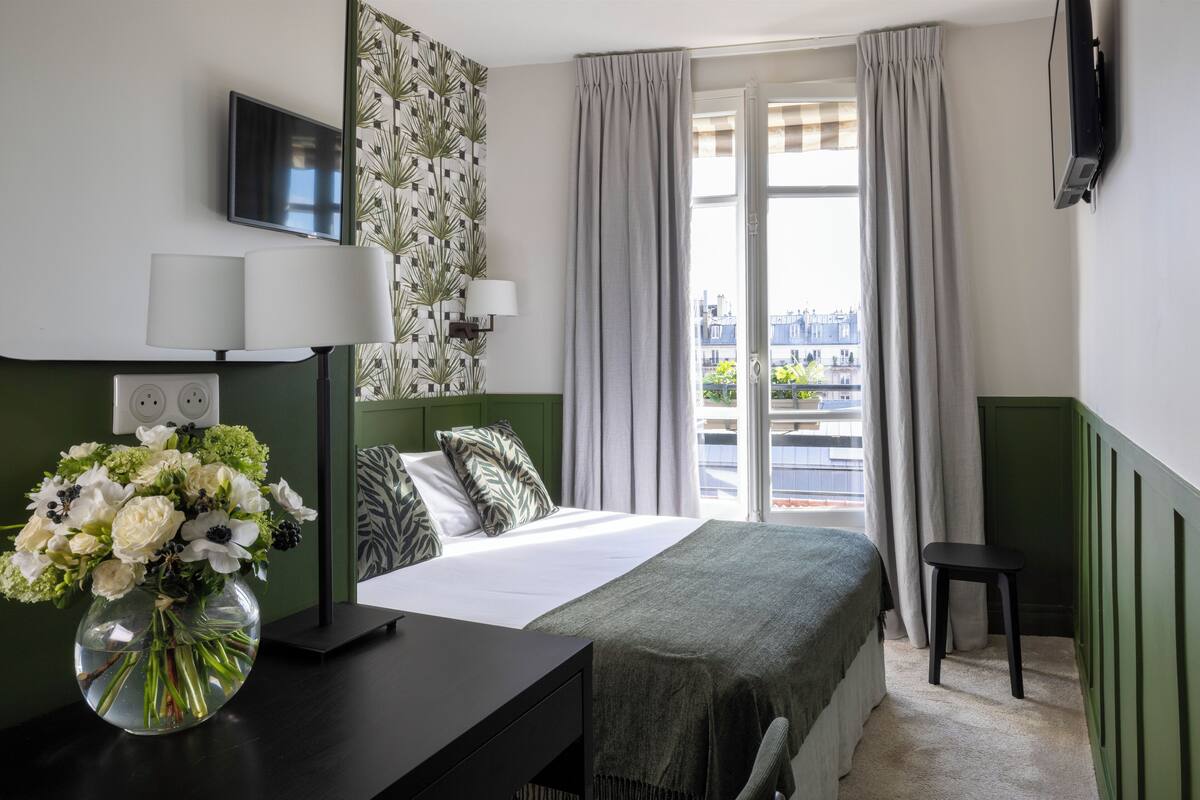 A hotel room with a bed, a desk with a lamp, a TV, a vase of flowers, and a view of the city through the window.