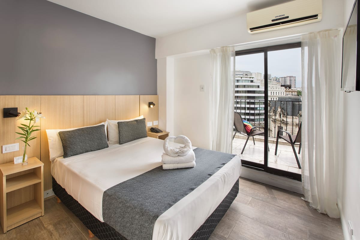 Habitación de hotel con una cama grande, una mesita de noche, una lámpara, una ventana con cortinas y vistas a edificios.