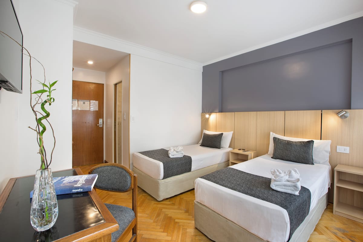 Habitación de hotel con dos camas, un escritorio, una silla, un florero con planta y cabecera de madera.