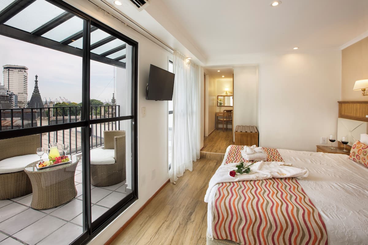 Una habitación de hotel con balcón, una cama con ropa de cama a rayas, un televisor montado en la pared y una zona de comedor con mesa y sillas.