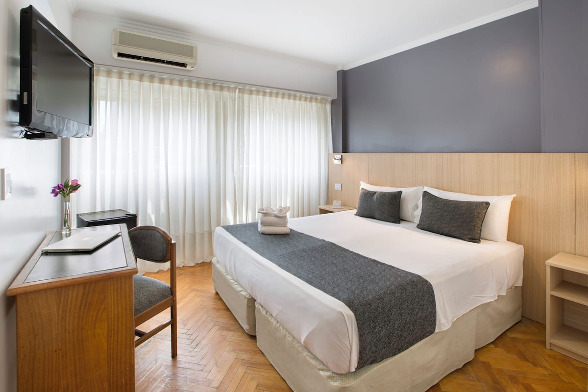 Habitación de hotel con cama, escritorio, silla, televisor y ventana con cortinas.