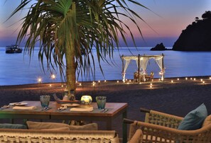 A beachside dining setup with a table and chairs, a palm tree, and a canopy structure with lights.