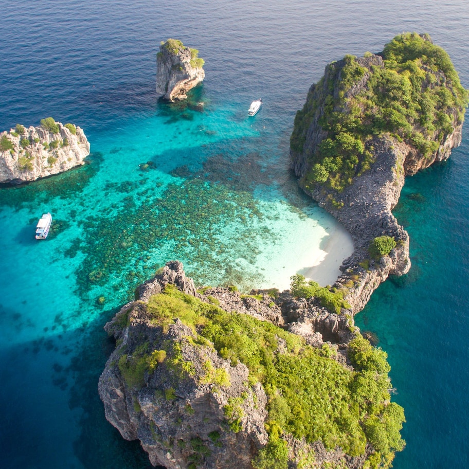 An aerial view of a coastal area with rocky formations, clear turquoise waters, and a sandy beach.