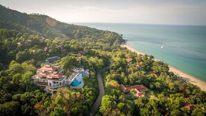 An aerial view of a resort with a swimming pool, beachfront, and lush greenery.