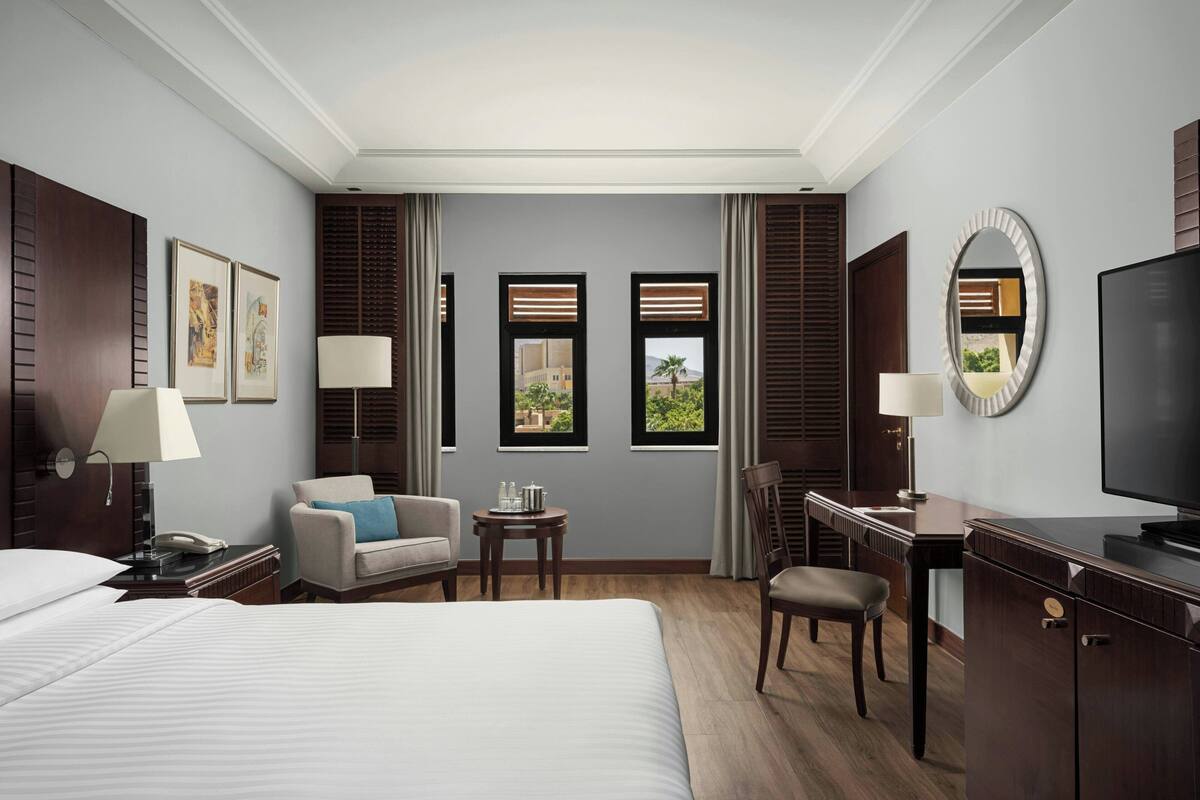 A hotel room with a large bed, a desk, a chair, a TV, and a view of the outdoors through windows.