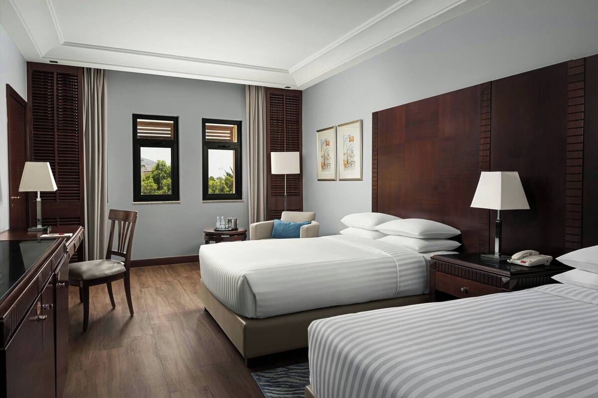 A hotel room with two beds, a desk, a chair, and a view of greenery through the windows.