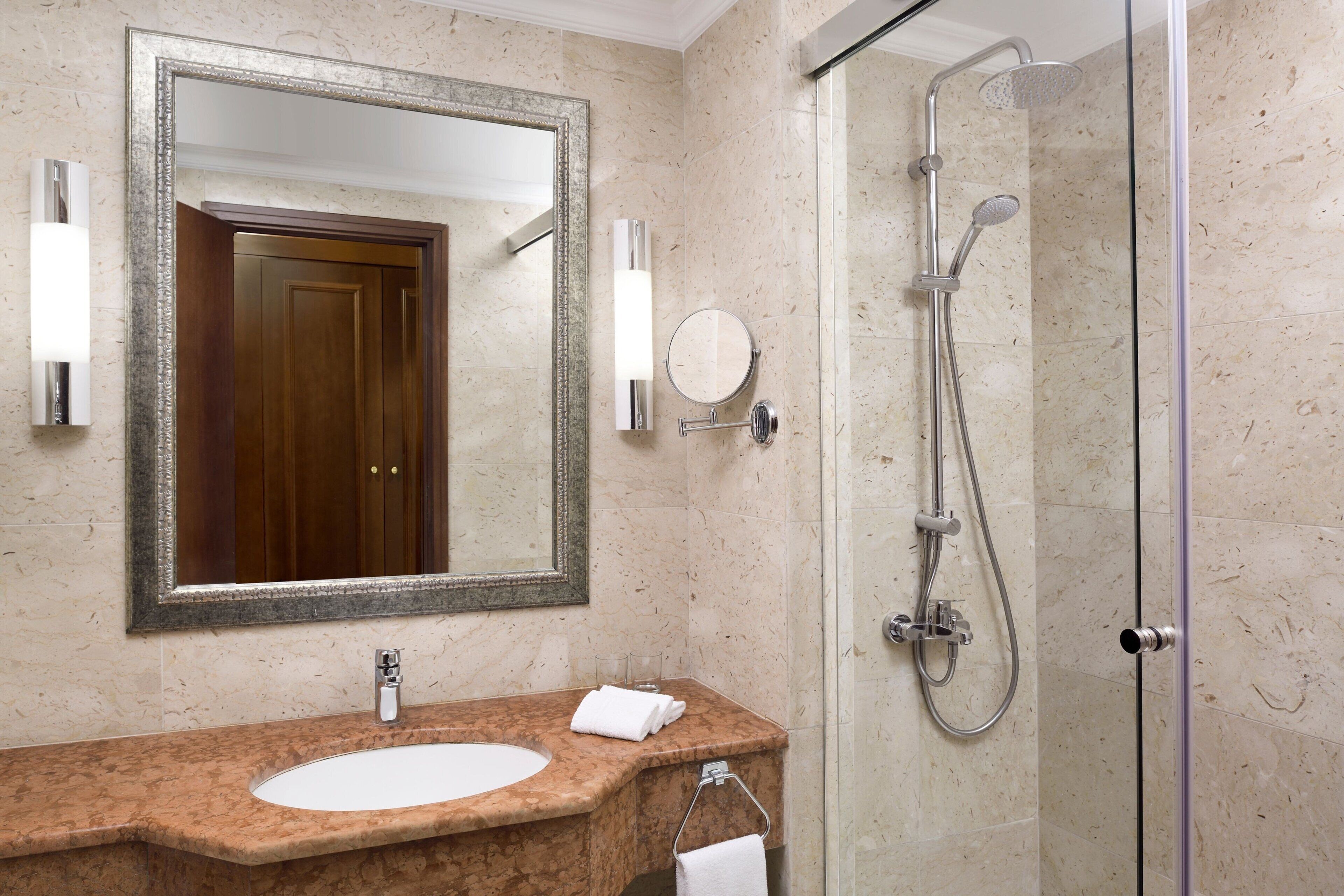 Un baño con lavamanos de mármol, un espejo grande, una ducha con paredes de vidrio y una puerta de madera.