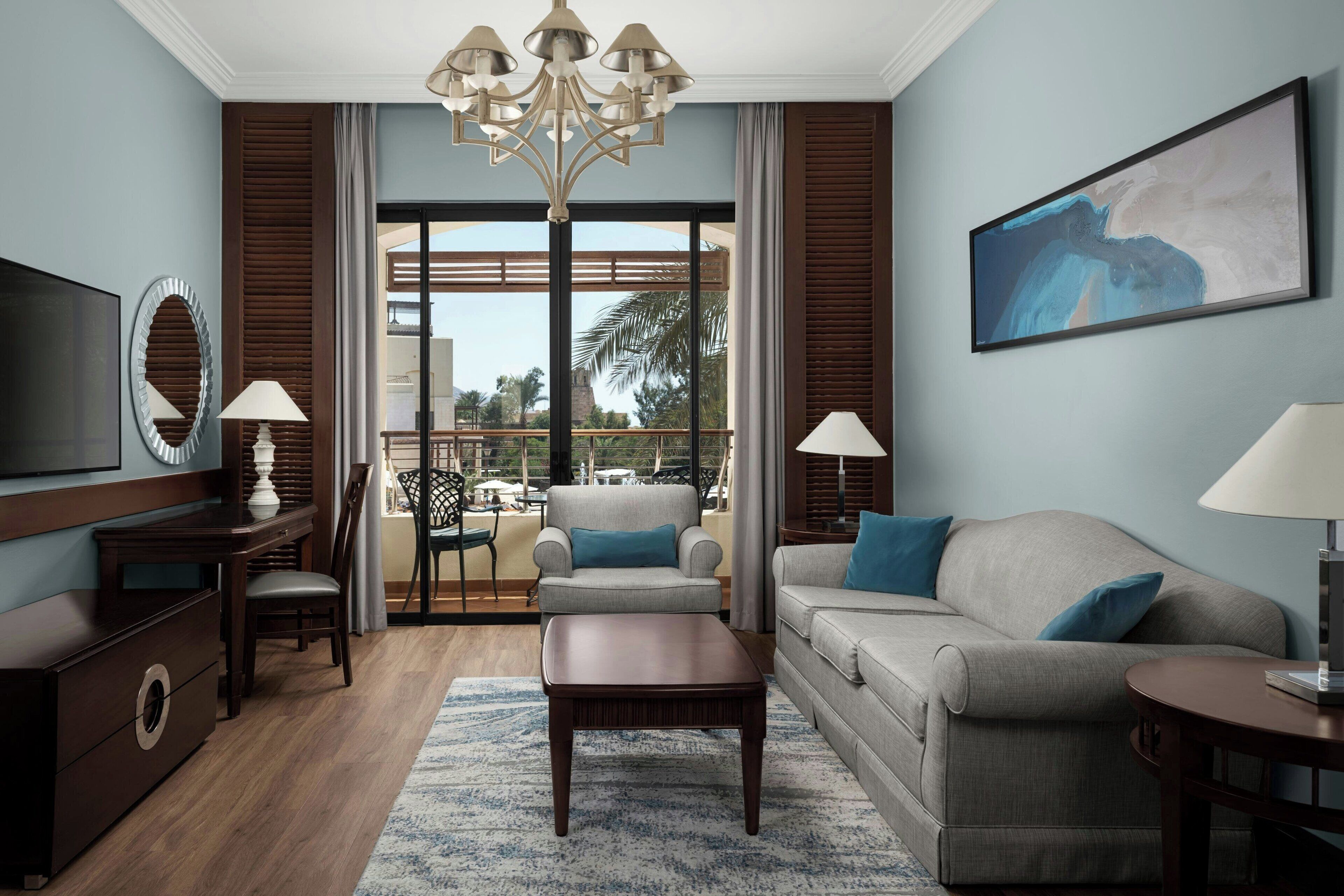 A modern living room with a sofa, armchair, coffee table, and TV. There is a balcony with outdoor furniture and a view of palm trees.