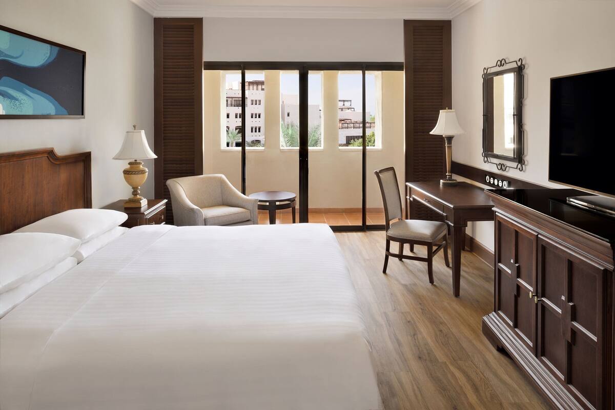 A hotel room with a large bed, a desk, a chair, a television, and a view of buildings through the windows.