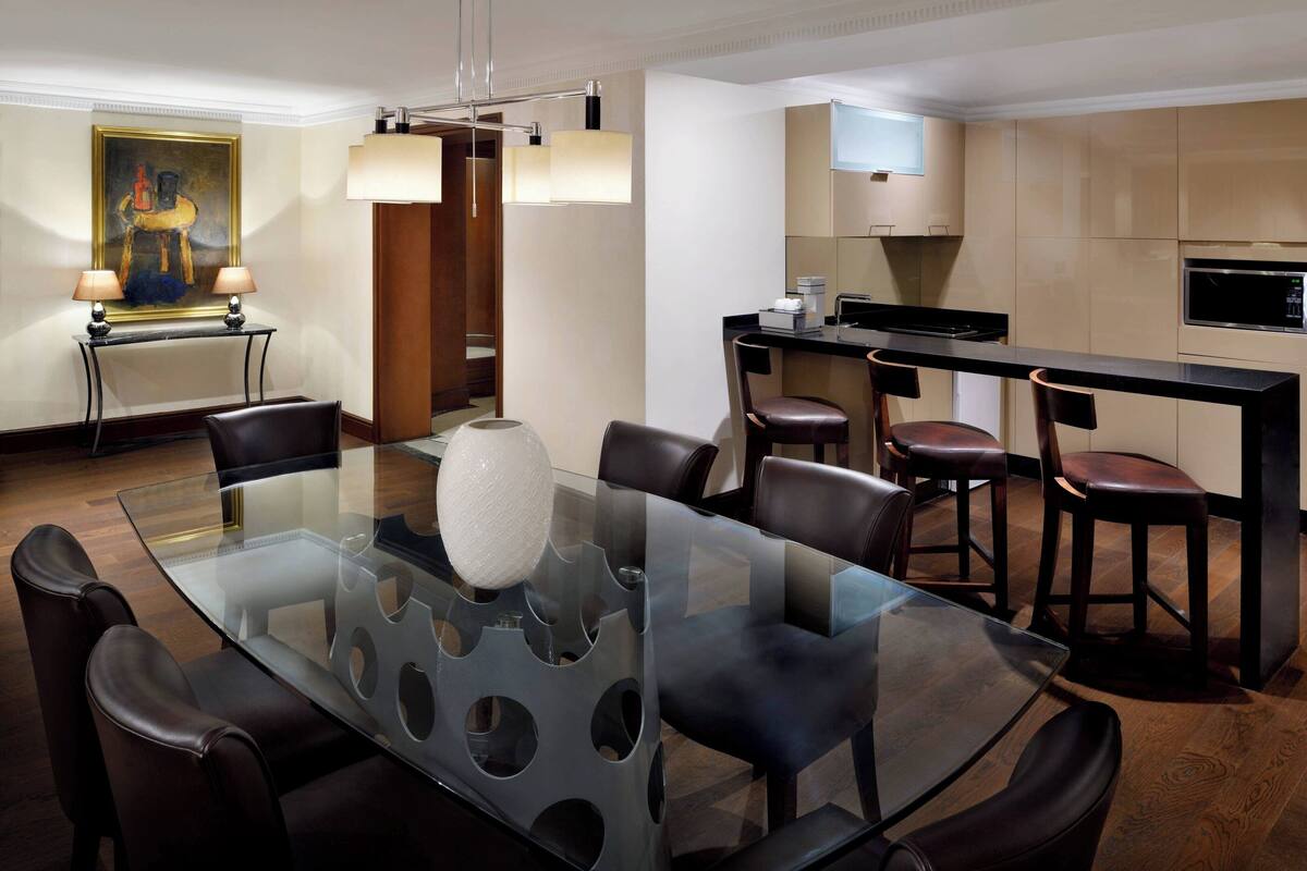 A modern dining area with a glass table, leather chairs, a painting on the wall, and a kitchen in the background.