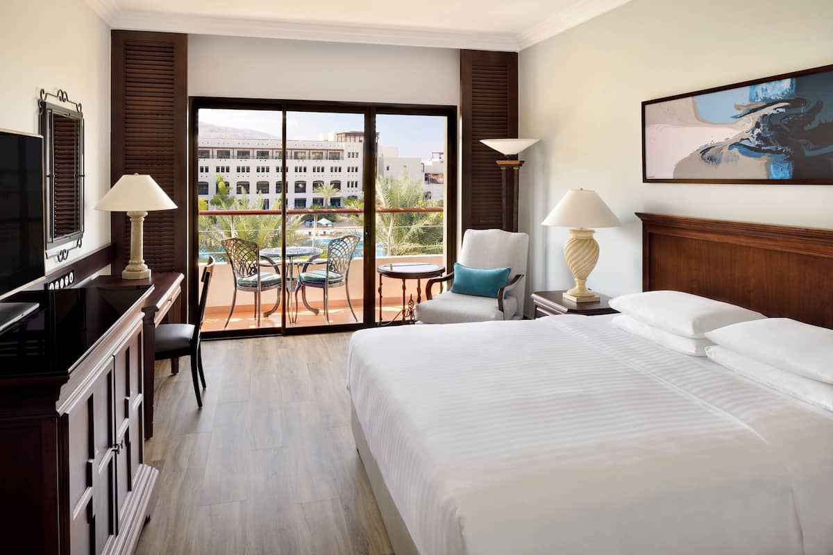 A hotel room with a large bed, a TV, a desk with a lamp, a chair, a balcony with a pool view, and a painting on the wall.