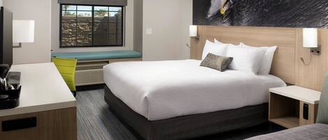 A hotel room with a large bed, a desk, a bench, and a window with a view of a rocky landscape.