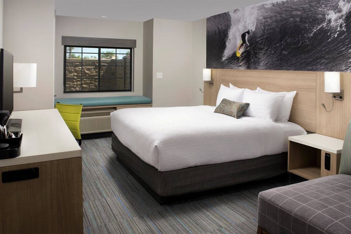 A hotel room with a bed, a desk, a chair, a bench, and a large window.