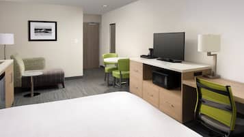 A hotel room with a bed, a desk with a TV, a chair, a small table, and a lamp.
