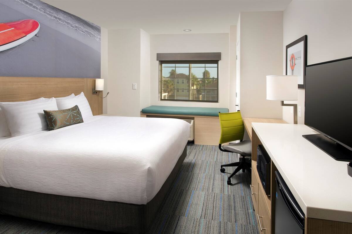 A hotel room with a large bed, a desk with a television, a window with a view of buildings, and a wall with a surfboard decal.