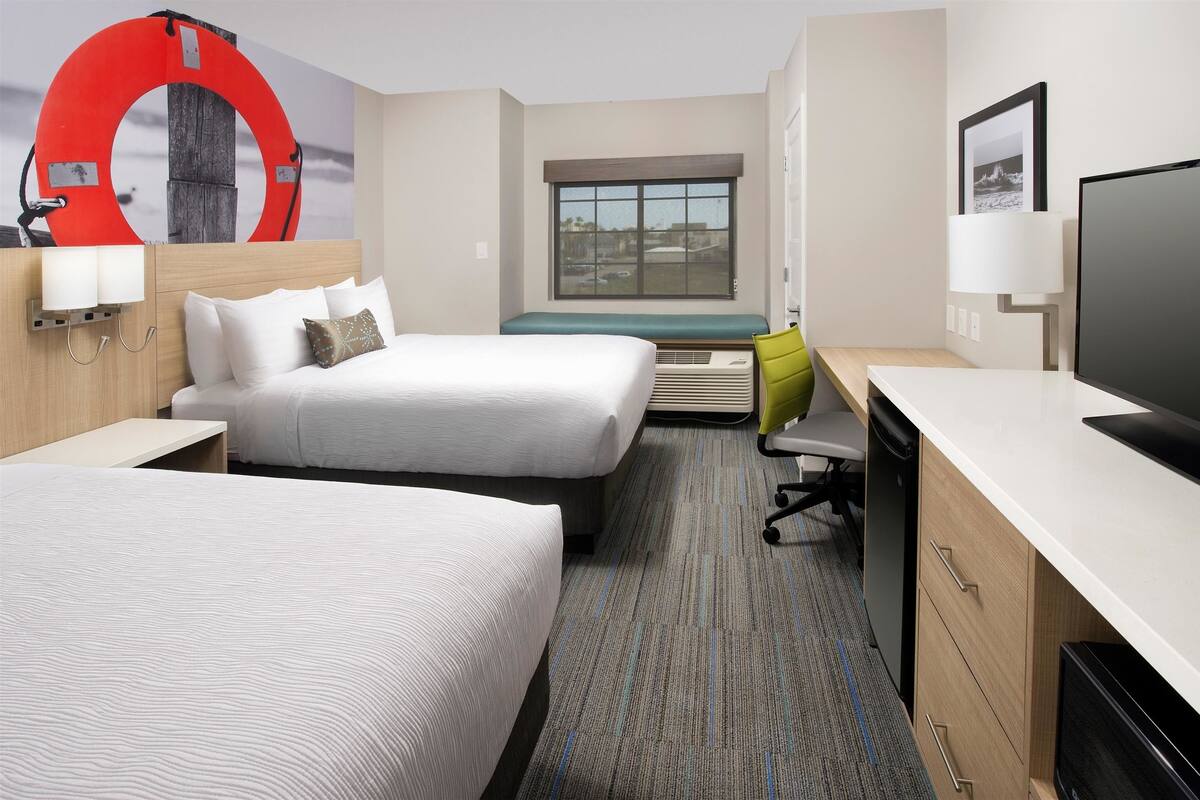 A hotel room with two beds, a desk, a TV, a window with a view, and a life preserver decoration on the wall.