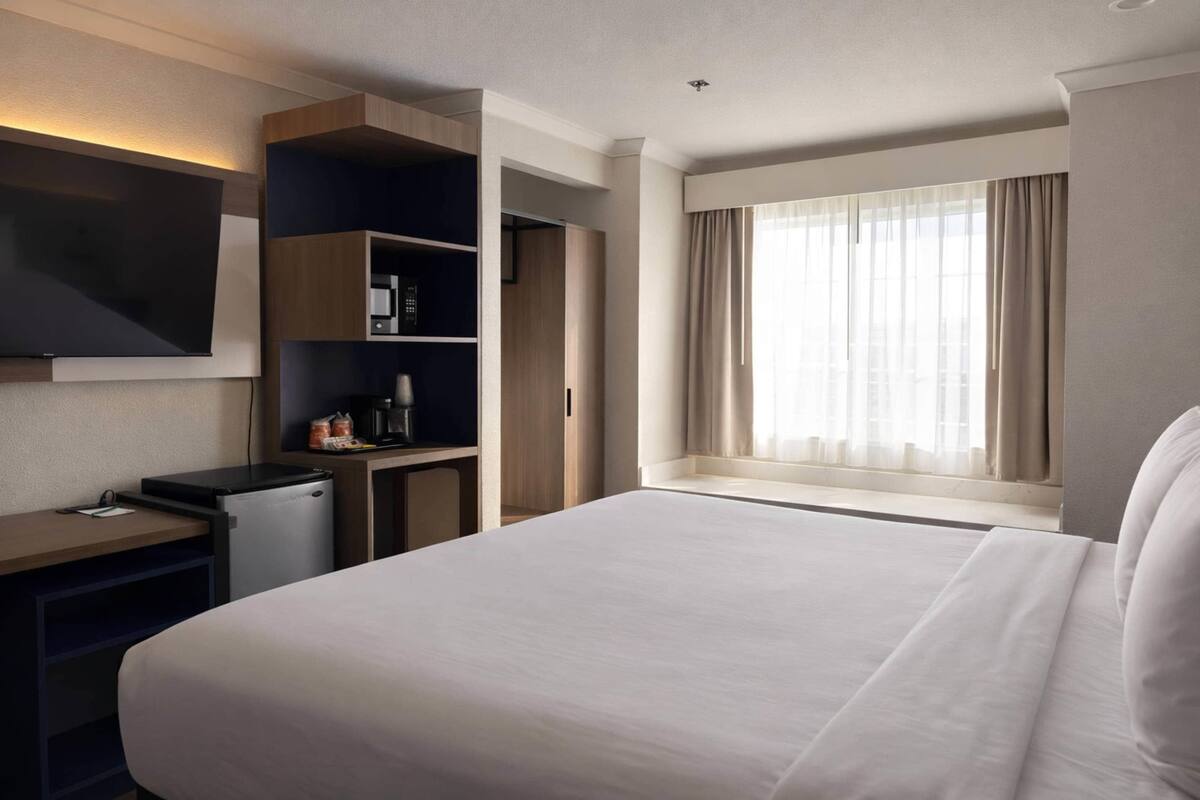 A hotel room with a large bed, a desk with a flat-screen TV, a mini-fridge, and a built-in shelving unit.