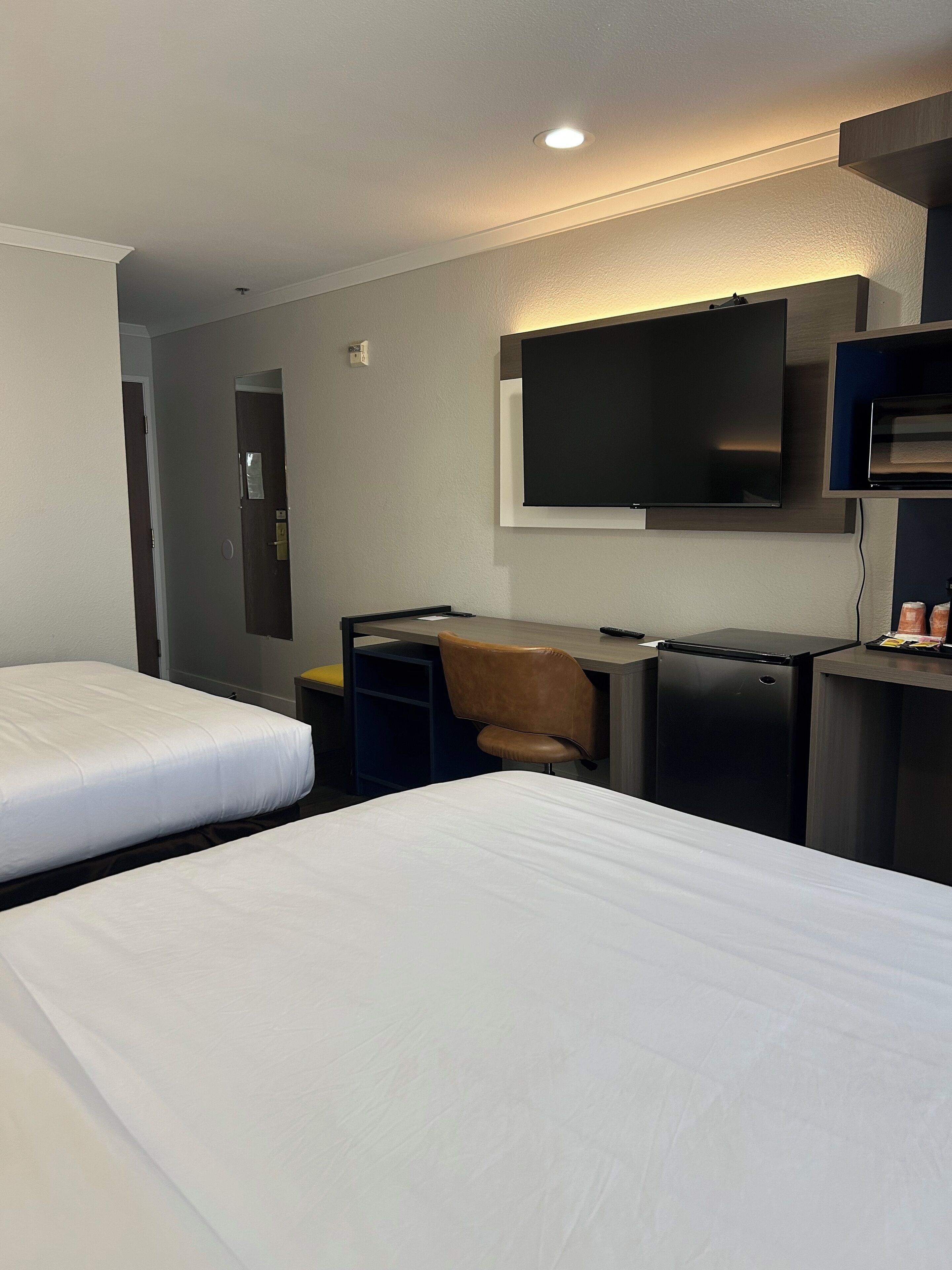 Habitación de hotel con dos camas individuales, un escritorio con silla, televisión en la pared y un refrigerador pequeño.