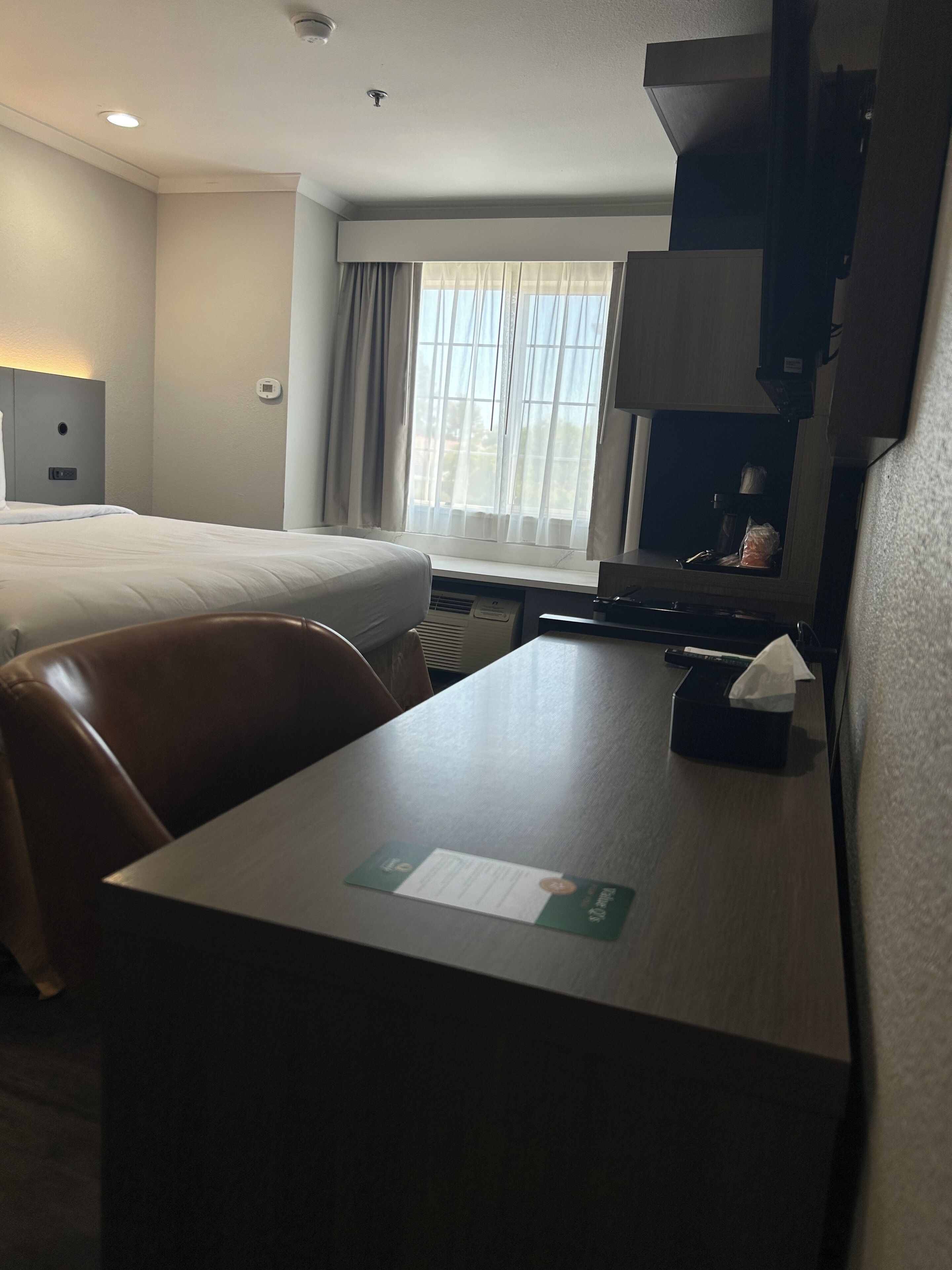 A hotel room with a bed, a desk, a chair, a window with curtains, a wall-mounted air conditioner, and a small table with a tissue box.